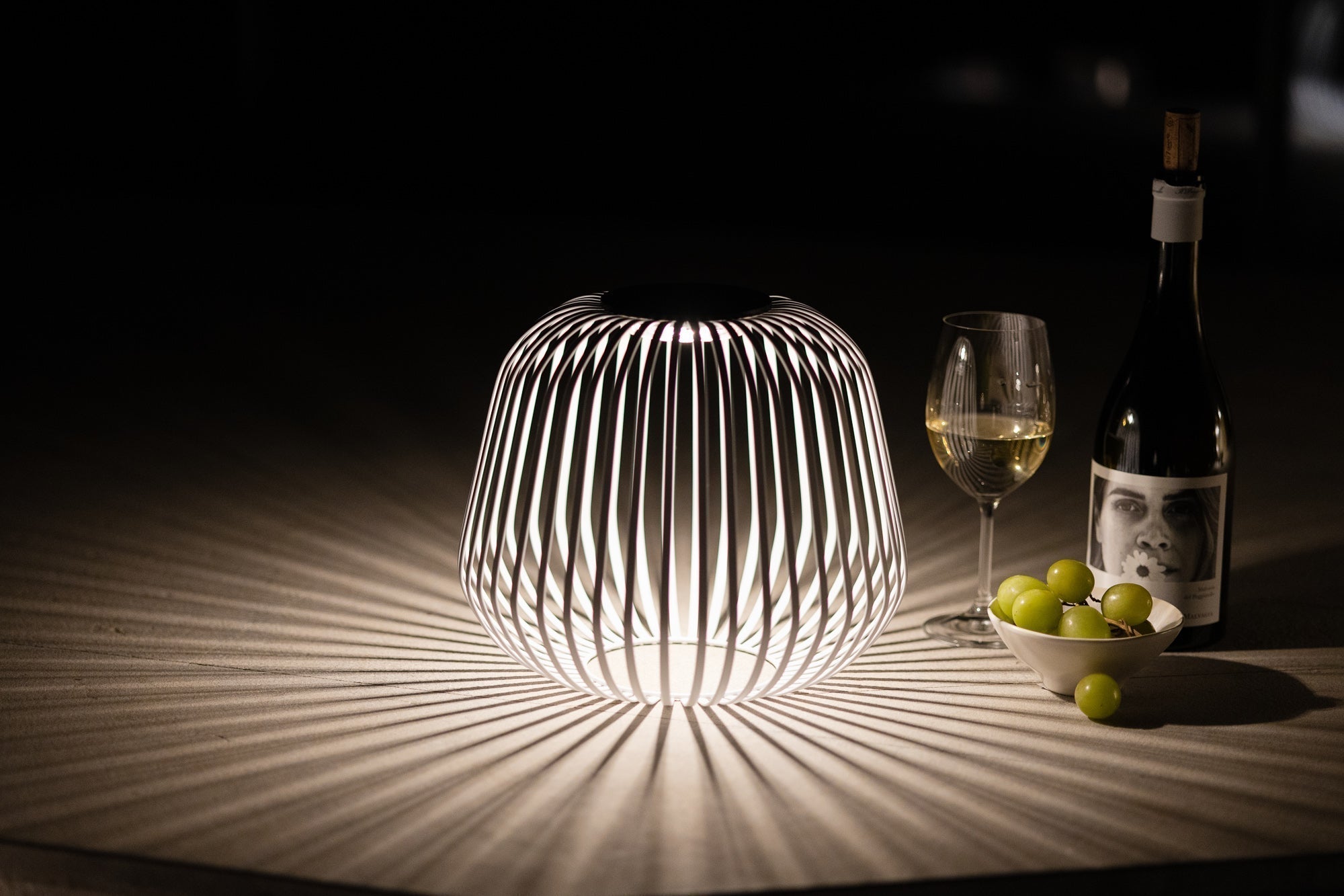 Die Solarlaterne Solis erzeugt ein dekoratives Licht- und Schattenspiel auf dem Tisch, daneben stehen ein Glas Weißwein, eine Weinflasche und eine Schale mit Trauben – ideal für stilvolle Abendmomente im Innen- oder Außenbereich.
