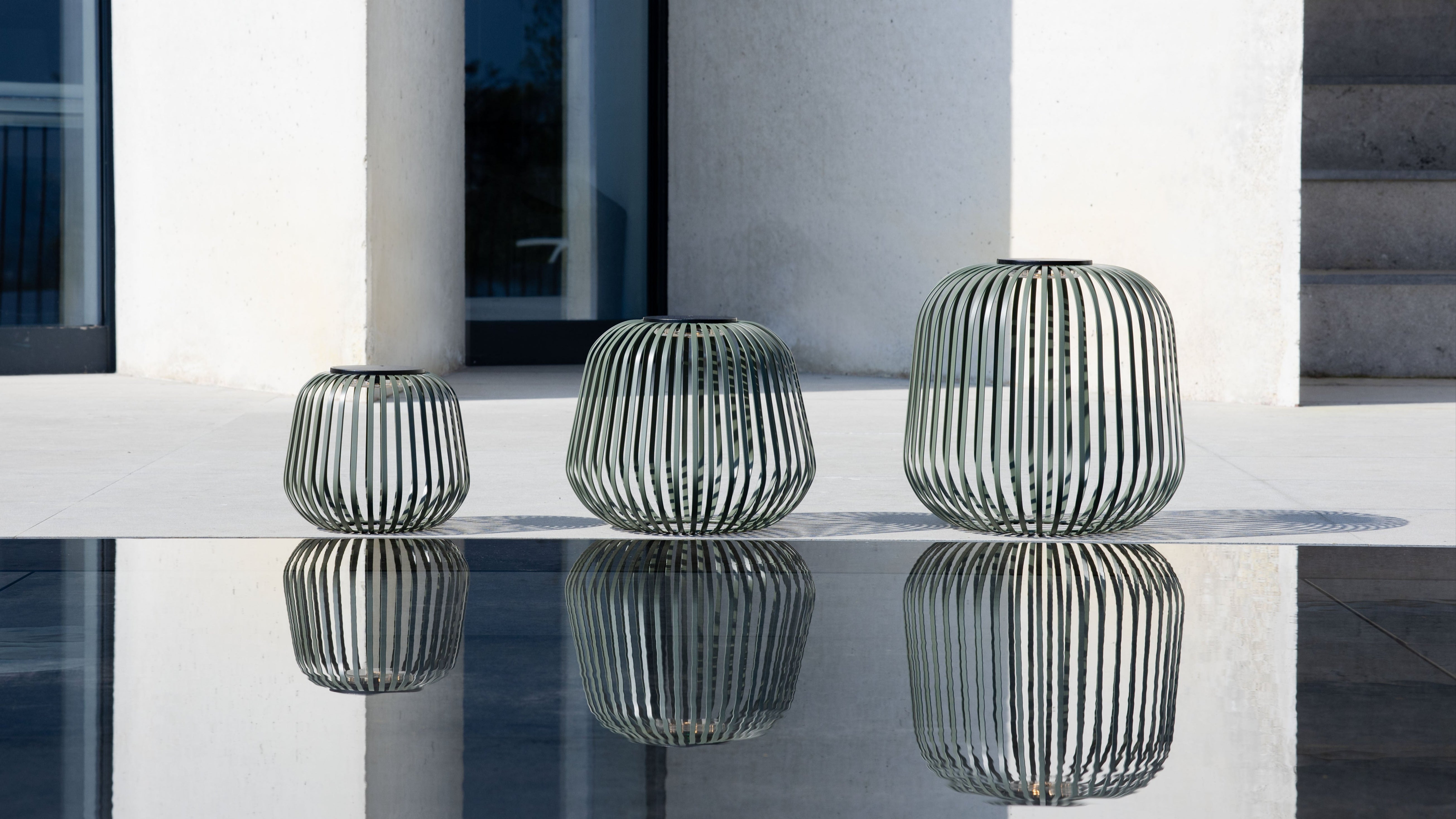 Drei grüne Solarlaternen in unterschiedlichen Größen reflektieren sich in einer Wasserfläche vor moderner Architektur – ideal für stilvolle, netzunabhängige Außenbeleuchtung im Garten oder auf der Terrasse