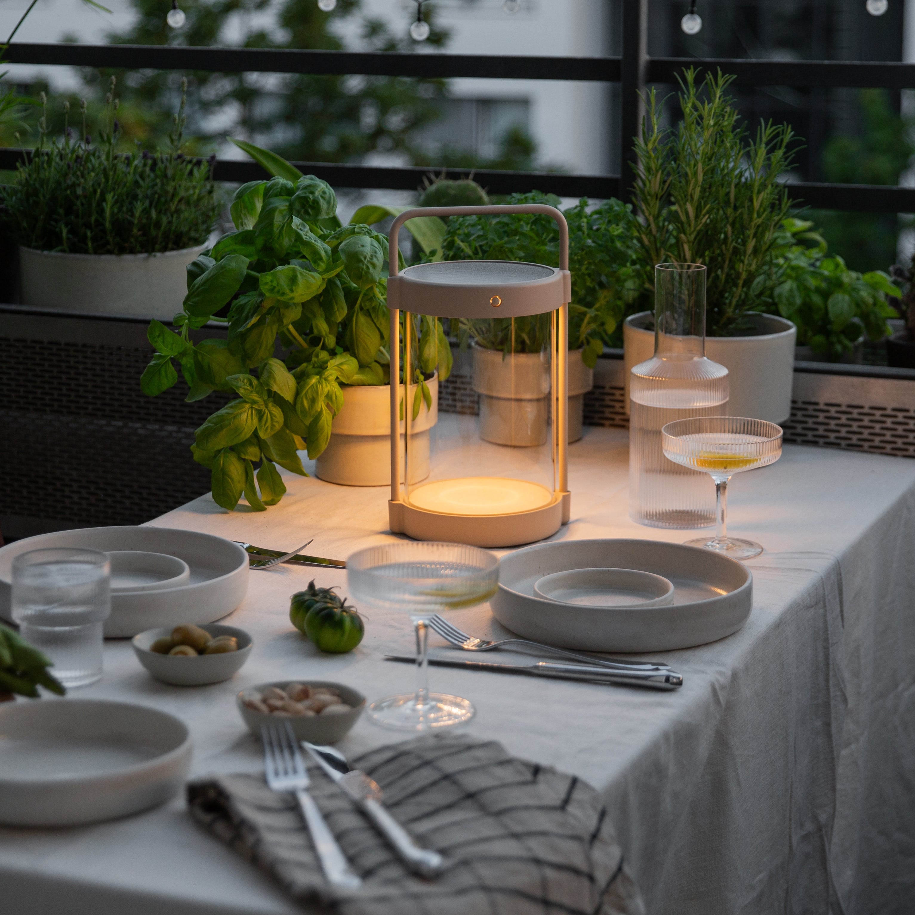 Beige LED Solarlampe von LUUK Studio auf gedecktem Tisch bei Dämmerung, stimmungsvolle Balkonbeleuchtung mit Kräutern im Hintergrund