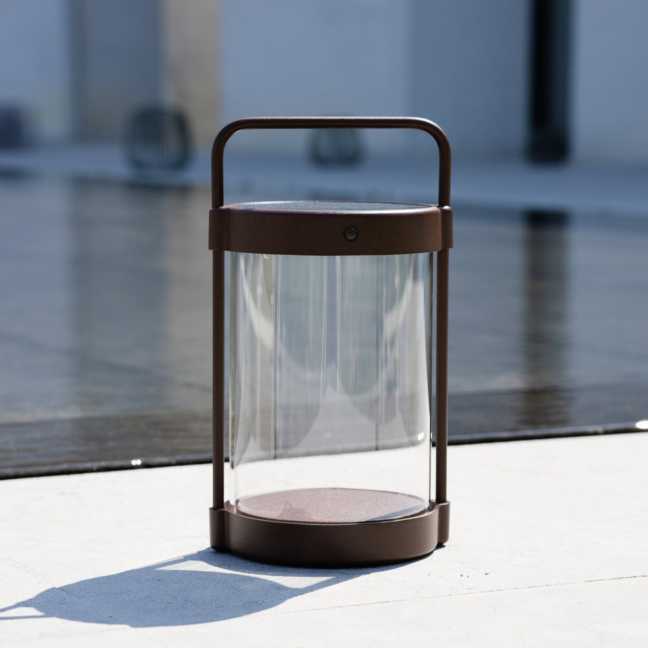 Caia Solarlaterne in Braun mit Glas im Tageslicht auf Terrasse – kabelloses Design-Leuchtobjekt für den Außenbereich mit Solaraufladung vor Naturpool