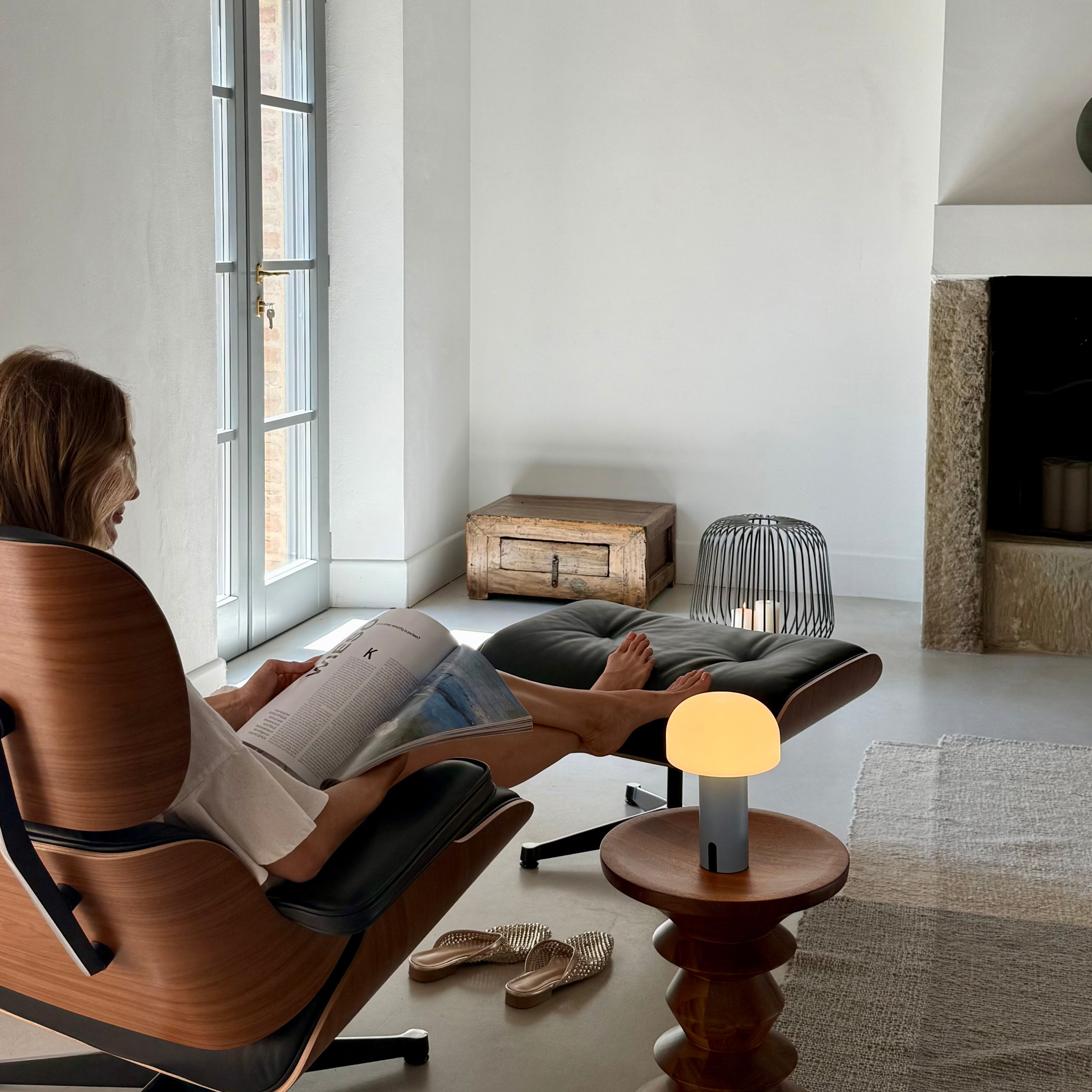 Eine Frau sitzt in einem Sessel im Wohnzimmer mit Kamin und liest ein Magazine. Auf dem Beistelltisch steht eine NOA Lampe in hellblau und eine SOLIS Solarlampe in XL und schwarz neben dem Kamin auf dem Fussboden.