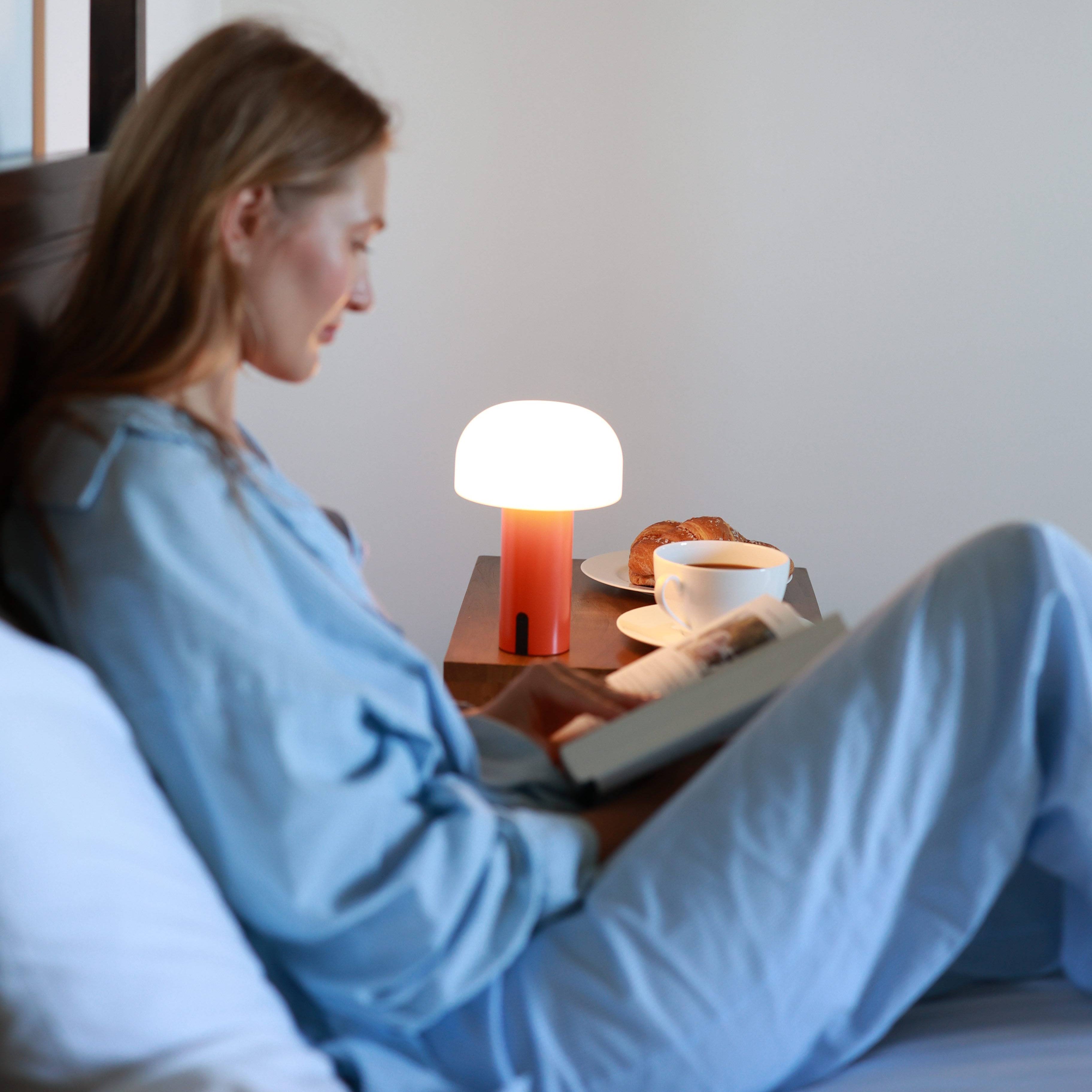Die mobile und kabellose NOA LED Laterne in einer entspannten Atmosphäre im Schlafzimmer mit Croissant, Kaffeetasse und einem Frau im Schalfanzug die ein Buch liest