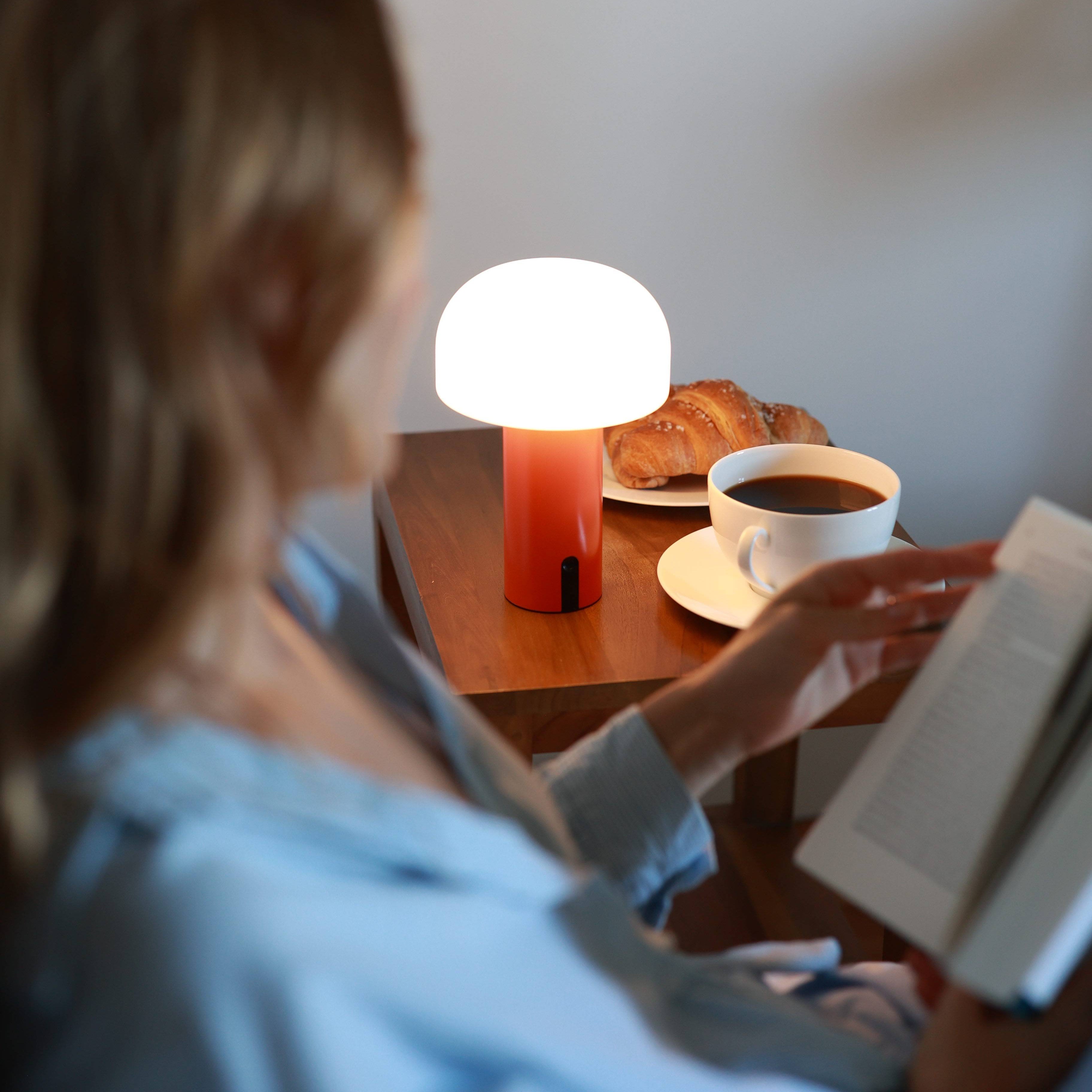 die minimalistische NOA Lampe in rot auf einem Nachtisch mit Croissant und Kaffeetasse sowie einer Frau im Schlafanzug die ein Buchliest