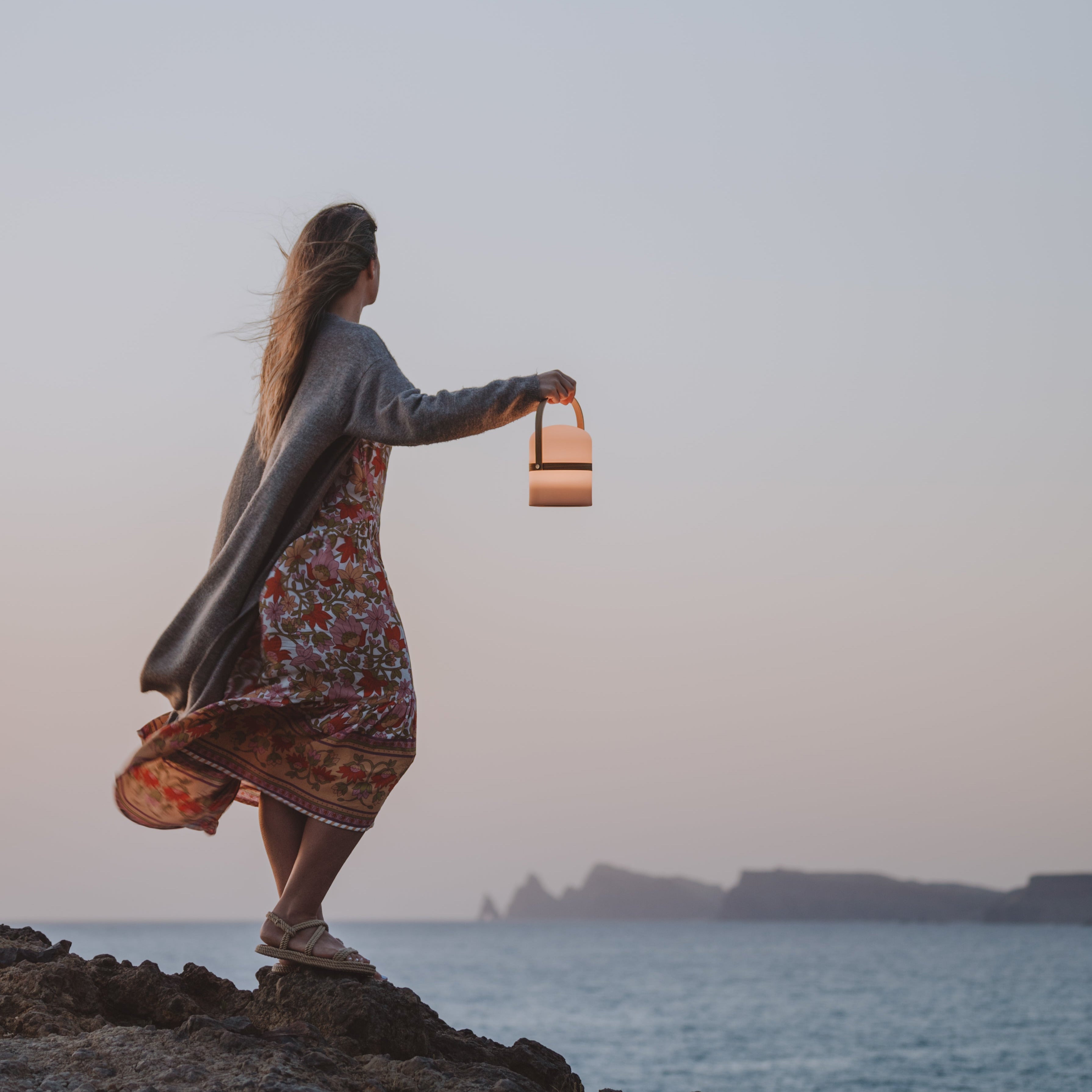 Frau mit tragbarer LED-Leuchte Luma bei Sonnenuntergang am Meer – stimmungsvolle Outdoor-Beleuchtung mit Designanspruch
