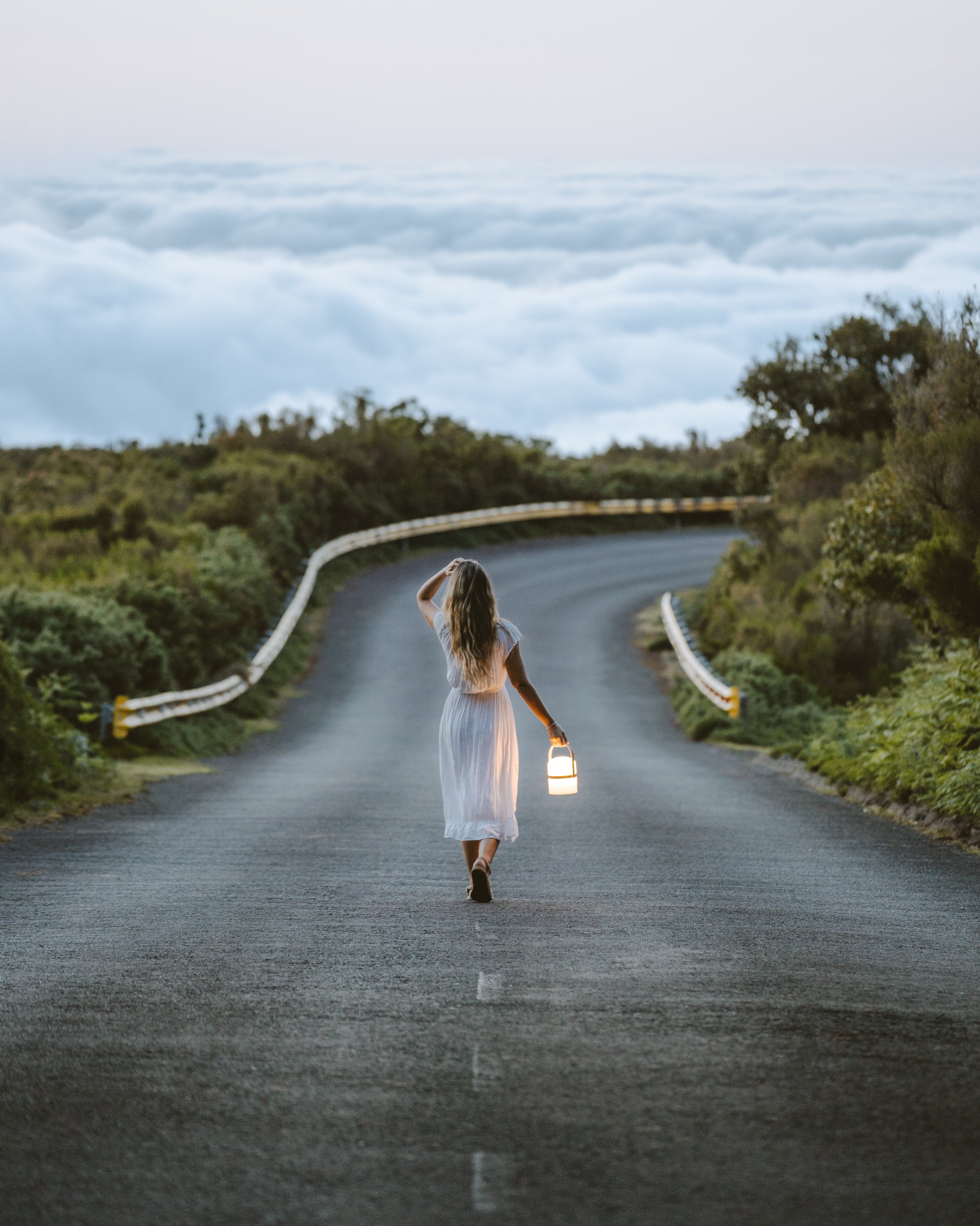Eine Frau läuft auf mediterraner Insel mit der mobilen Luma LED Laterne von LUUK Studio in der Hand auf einer Küstenstraße in der Dämmerung