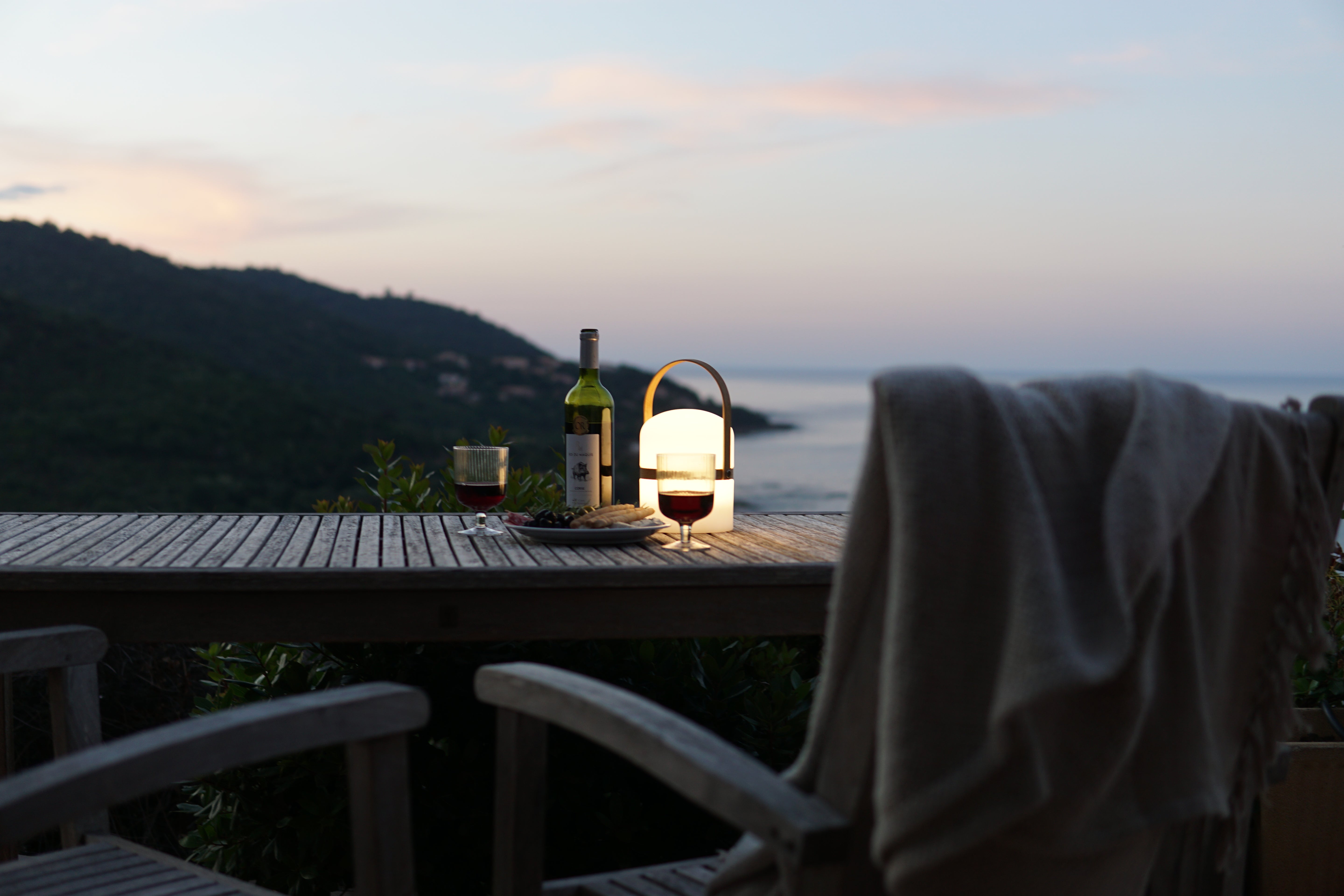 Die Luma Tischleuchte von LUUK Studio auf Terrasse mit Holztisch in der Abendstimmung mit Wein, Antipasti und Blick übers Meer