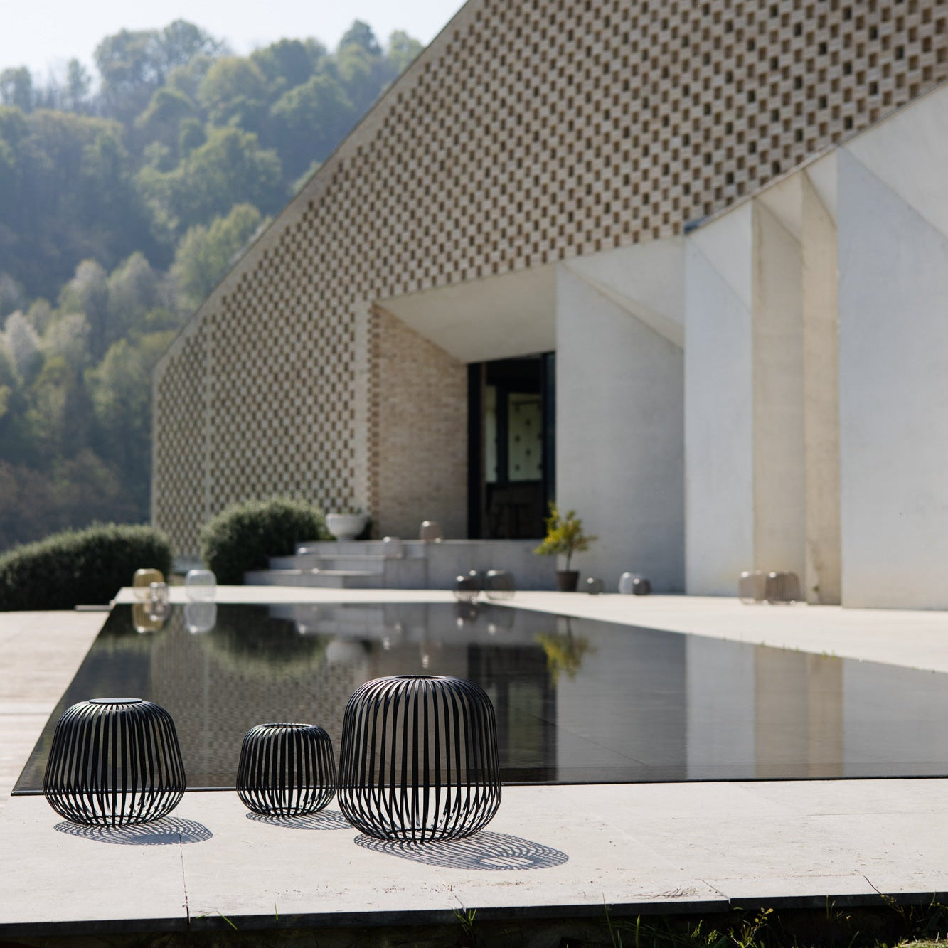Drei schwarze Outdoor-Leuchten von LUUK Studio mit Schattenwurf auf moderner Steinterrasse vor architektonischer Fassade und Naturpool Becken