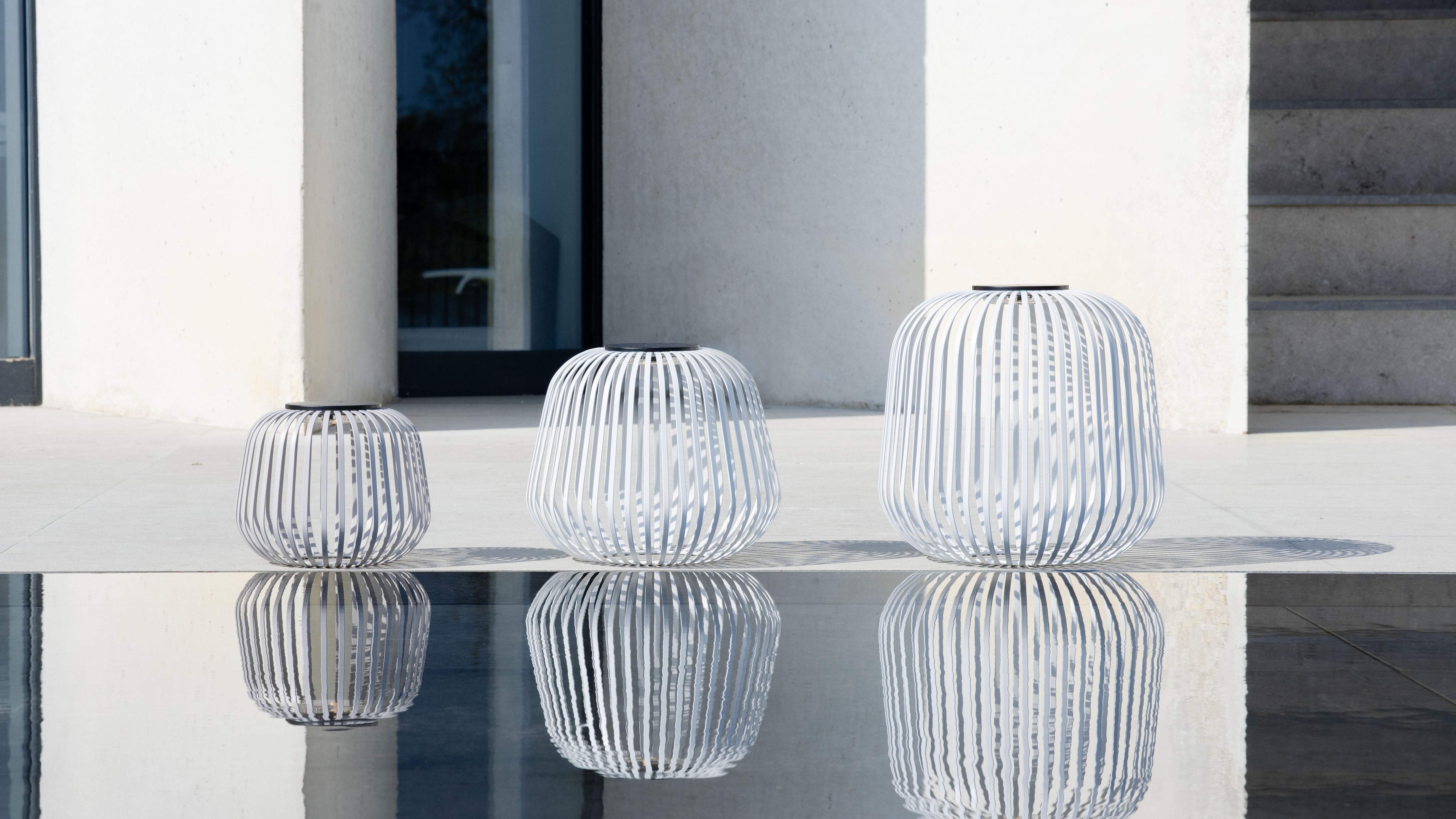 Drei Solis Solarlaternen in weiss auf moderner Terrasse – als dekorative Windlichter und stilvolle Kerzenhalter für stimmungsvolle Außenbeleuchtung mit eleganten Lichtreflexionen im Naturpool.