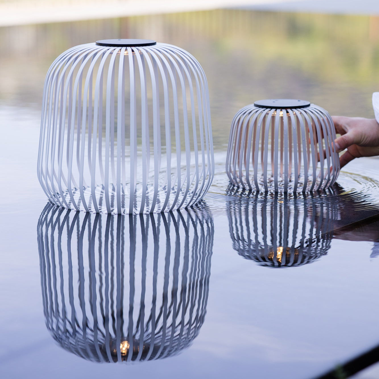 Zwei weiße Solis Outdoor Solarlaternen mit mobilem Solarmodul auf Wasseroberfläche mit Spiegelung, eine wird von Hand abgesetzt