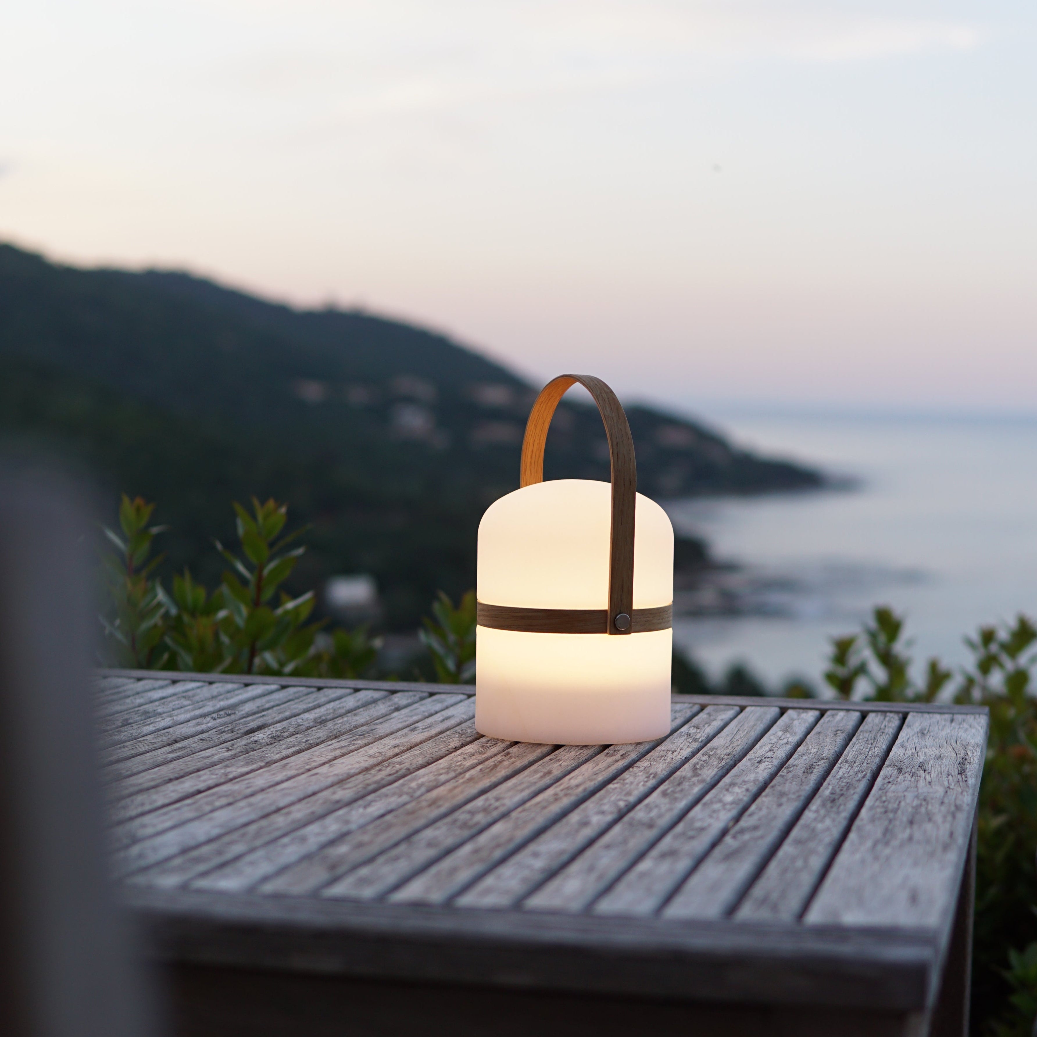 Luma Tischlaterne mit warmweißem Licht auf Terrassentisch in der Dämmerung mit Weitblick auf die Küste