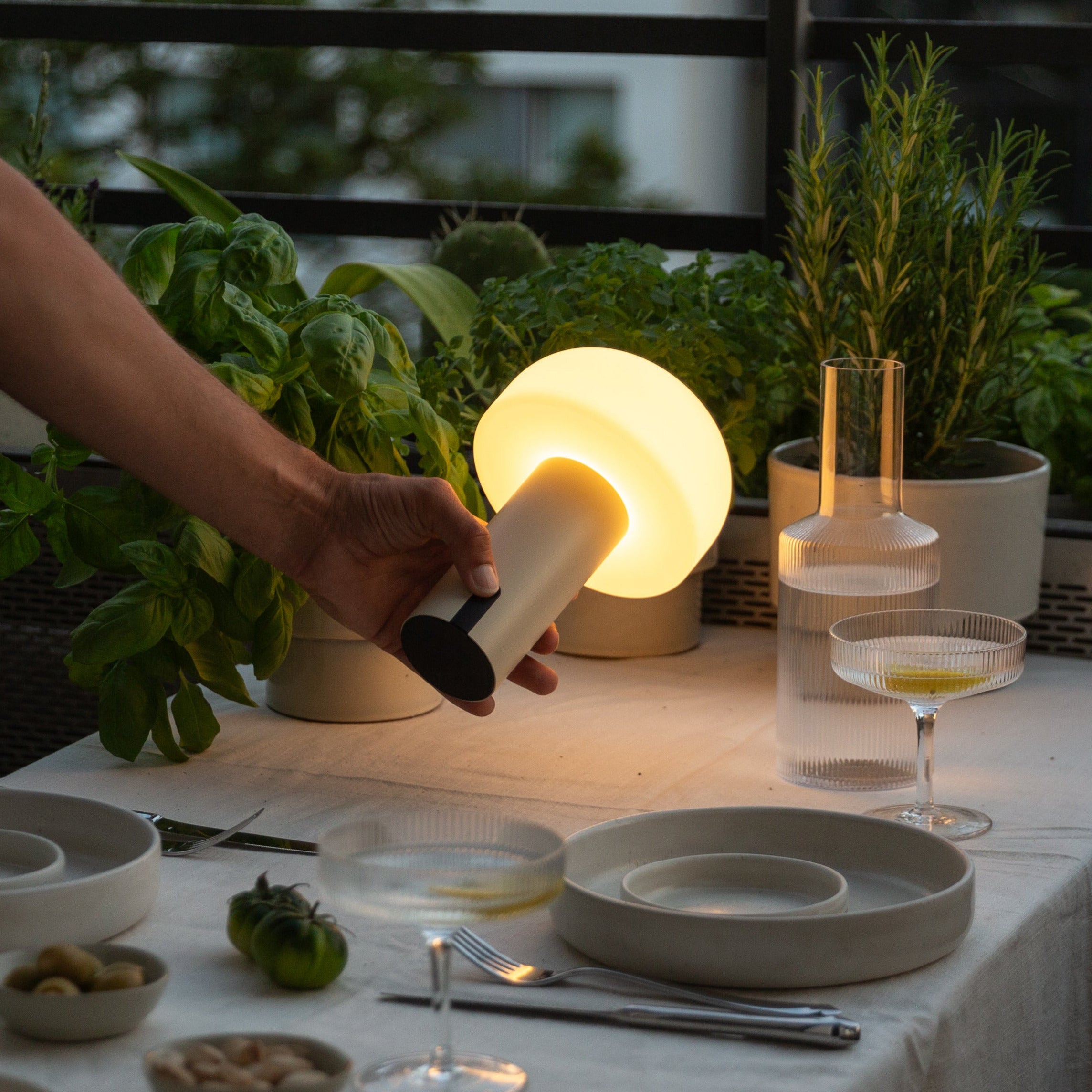 Kabellose Noa LED-Tischleuchte in hellem Beige auf einem gedeckten Esstisch im Outdoor-Bereich, dimmbares Licht für stimmungsvolle Abendstunden auf Balkon oder Terrasse.