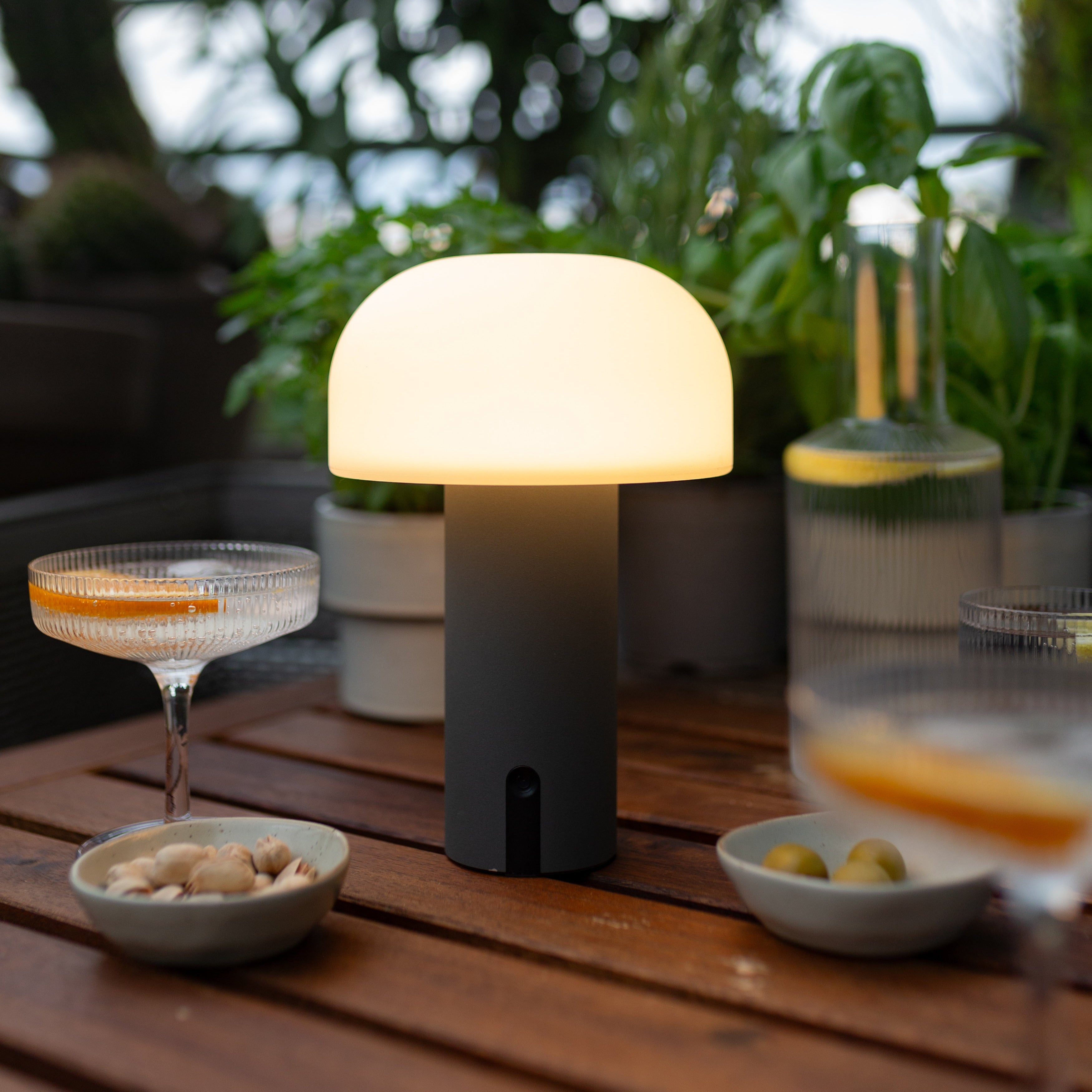 Mobile Noa LED-Pilzleuchte in grau spendet stimmungsvolles Licht auf gedecktem Terrassentisch – kabellose Tischlampe für Outdoor Dinner und Sommerabende