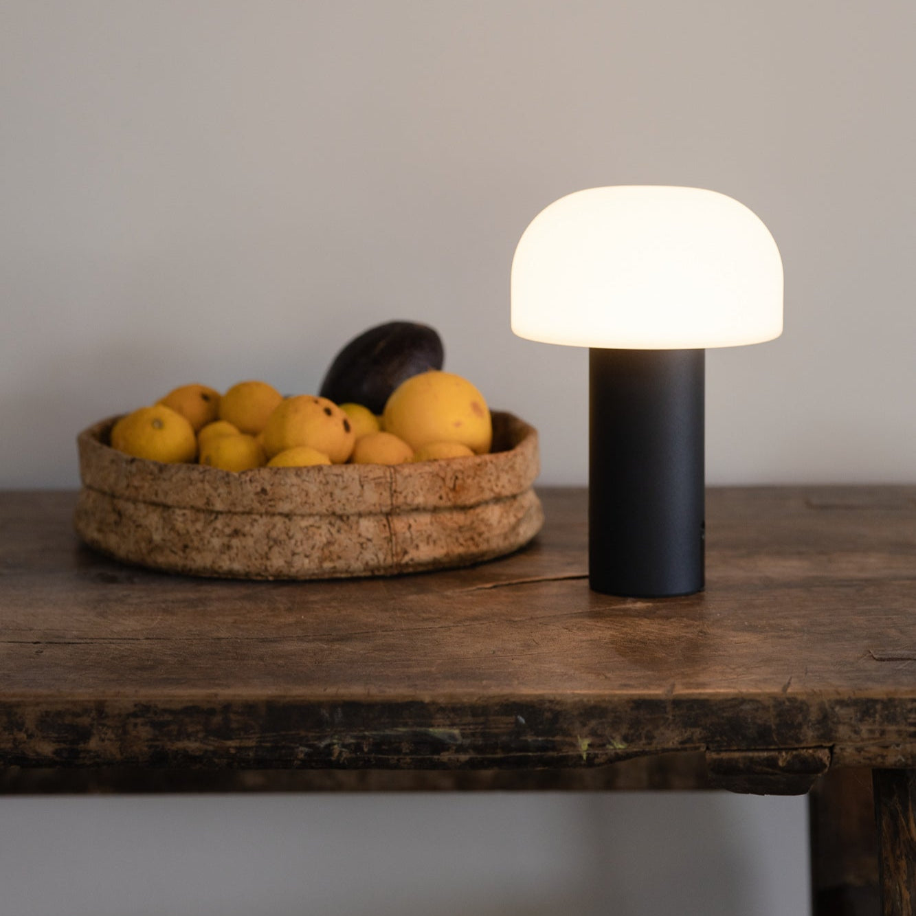 Noa LED Pilztischleuchte mit Akku von LUUK Studio in elegantem Schwarz auf einem rustikalen Holztisch – modernes Lichtobjekt mit weichem Schein für stilvolle Innenräume