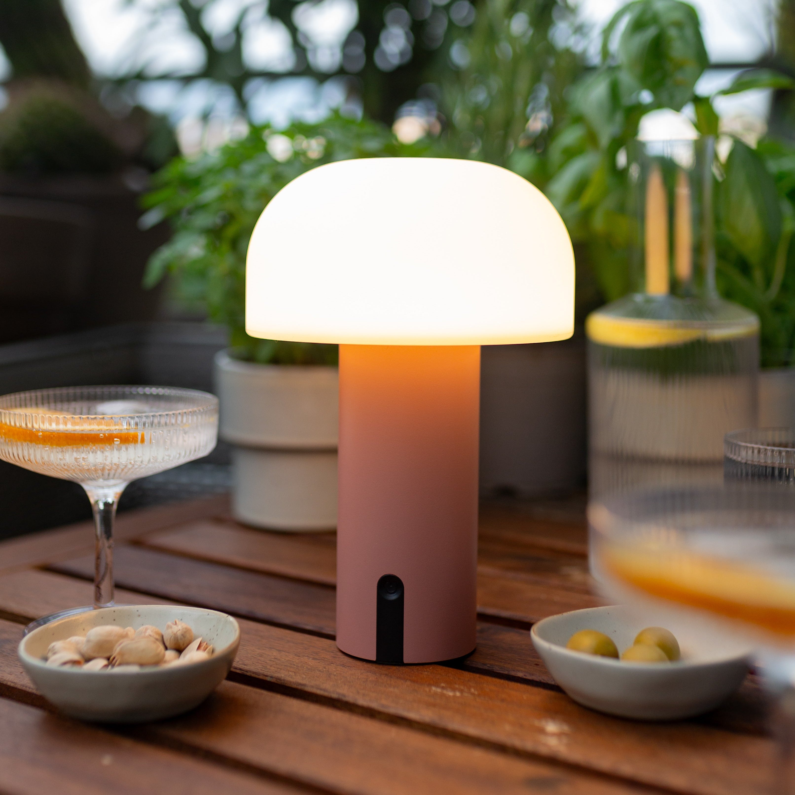 Noa Akku LED-Tischlampe ohne Kabel in Koralle-Terracotta auf gedecktem Terrassentisch mit Drinks und Kräutern im Hintergrund bei Abenddämmerung