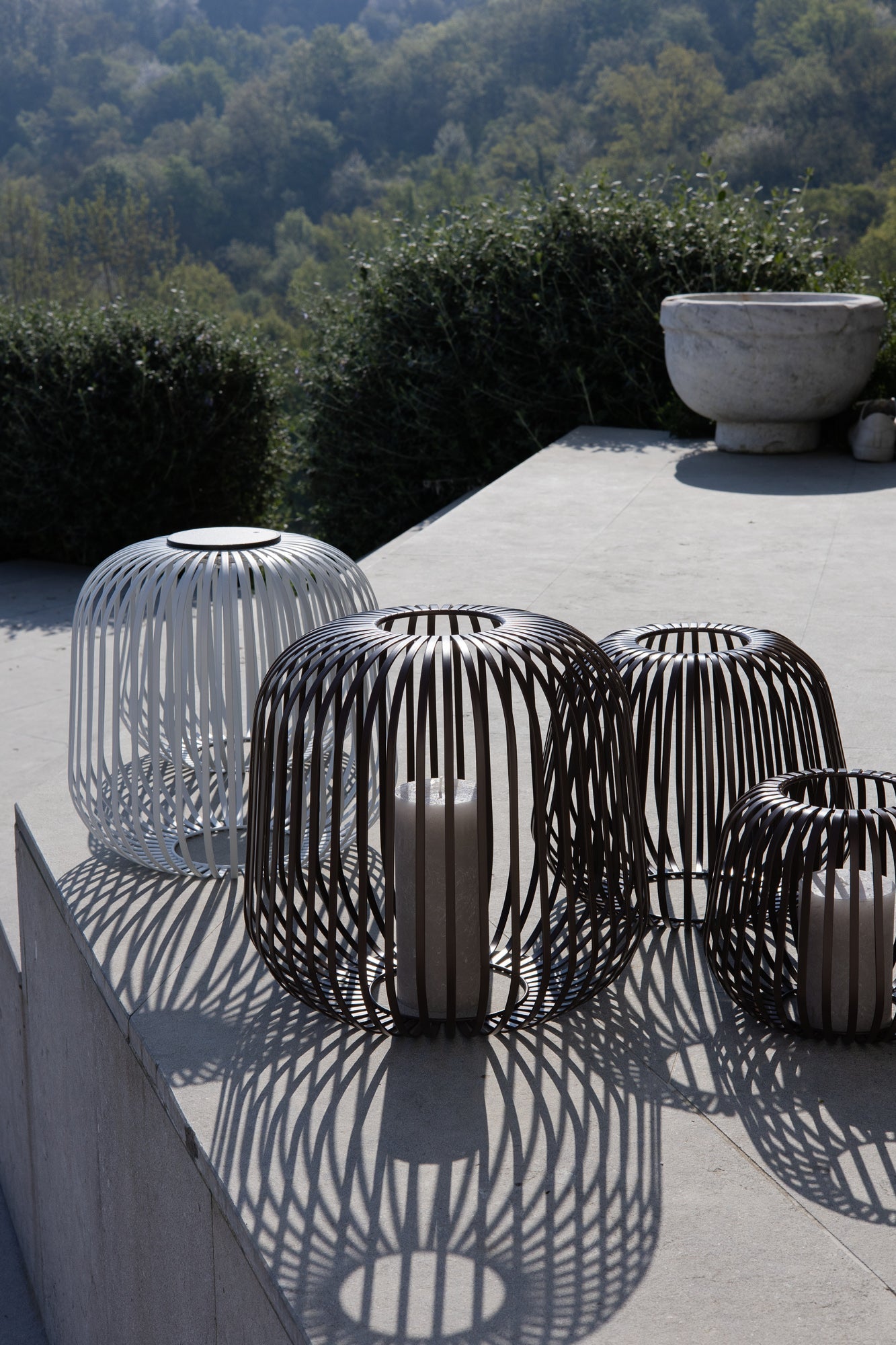 Vier LUUK Studio Outdoorleuchten aus Metall mit Solarmodul, Kerze und Schattenwurf auf sonniger Terrasse mit Landschaft im Hintergrund