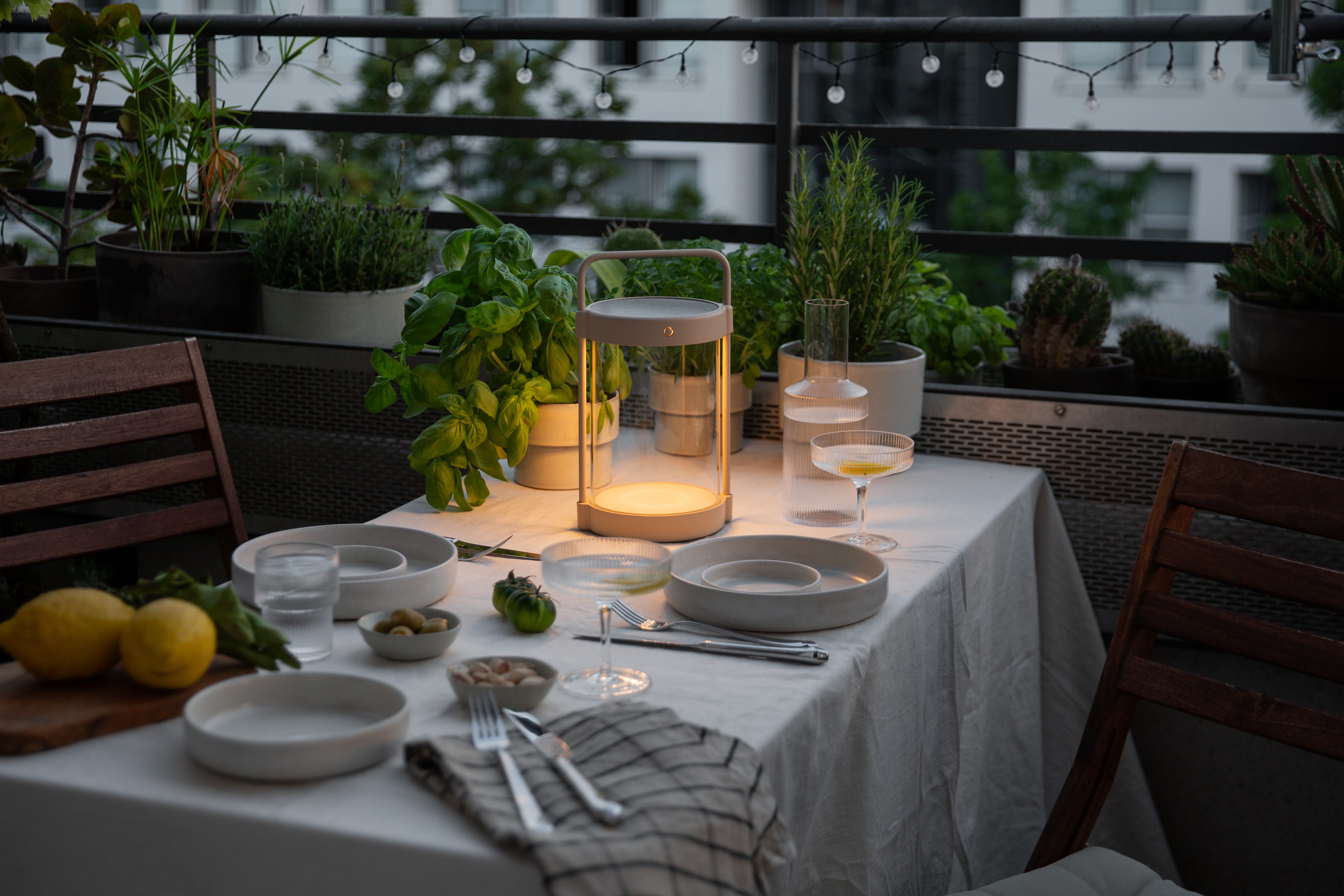 Die LED-Solarlaterne caia sorgt auf einem liebevoll gedeckten Balkontisch mit Kräutern, Geschirr und Getränken für stimmungsvolles Licht – ideal für laue Sommerabende und urbane Outdoor-Dinner.