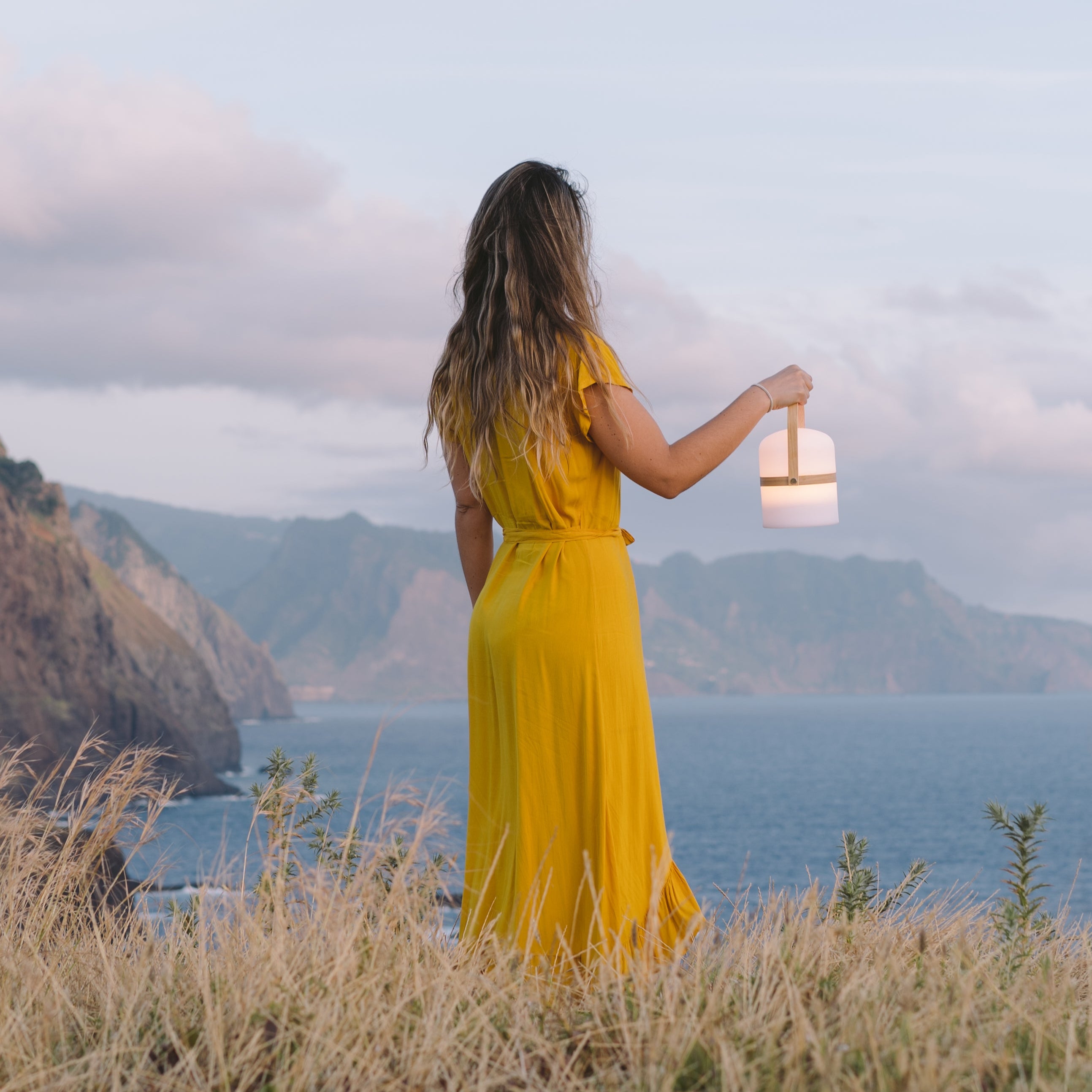 Frau hält gedimmte Luma Laterne in der Hand mit Blick übers Meer und genießt die Aussicht und die mediterrane Abendstimmung 