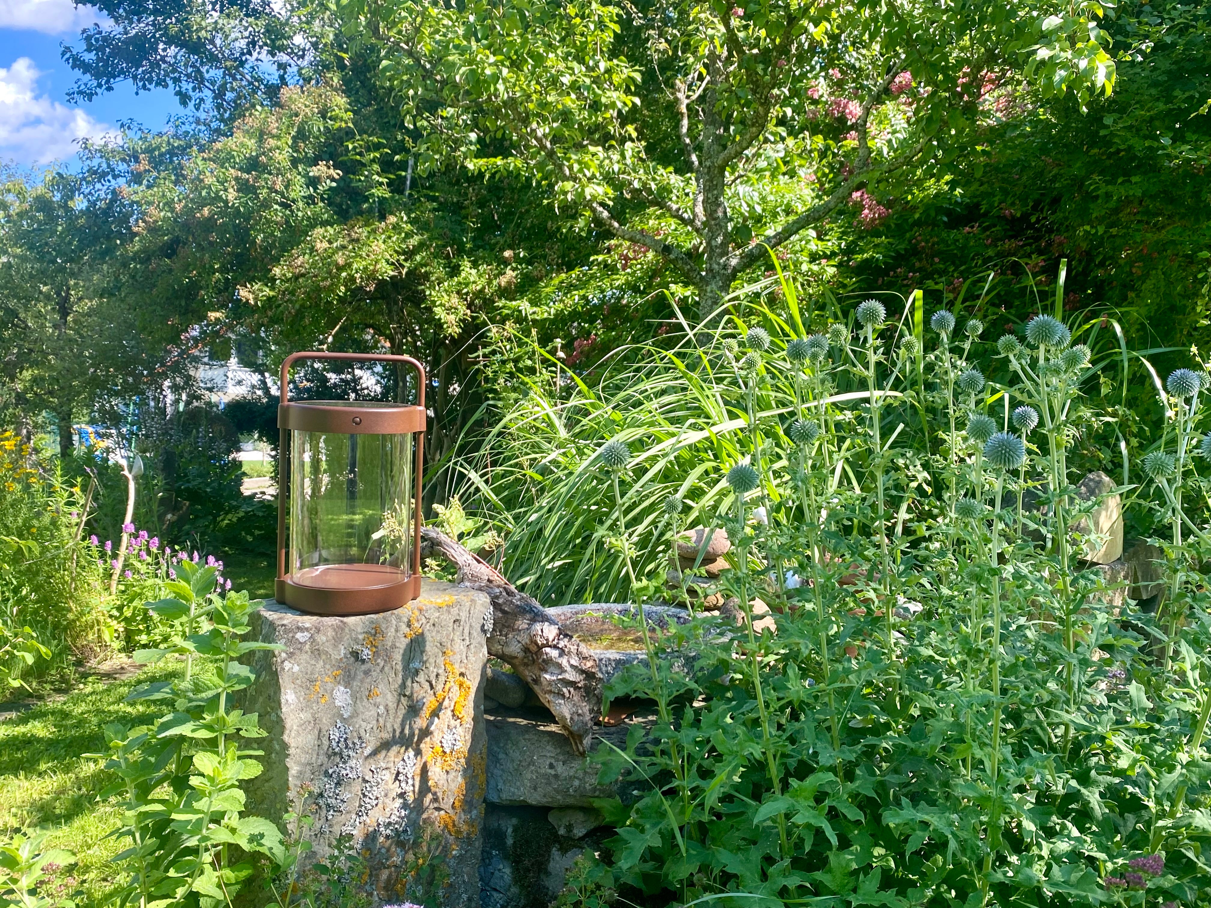Braune Solarlaterne Caia von LUUK Studio auf Naturstein-Sockel im blühenden Wildblumen Garten bei Sonnenschein
