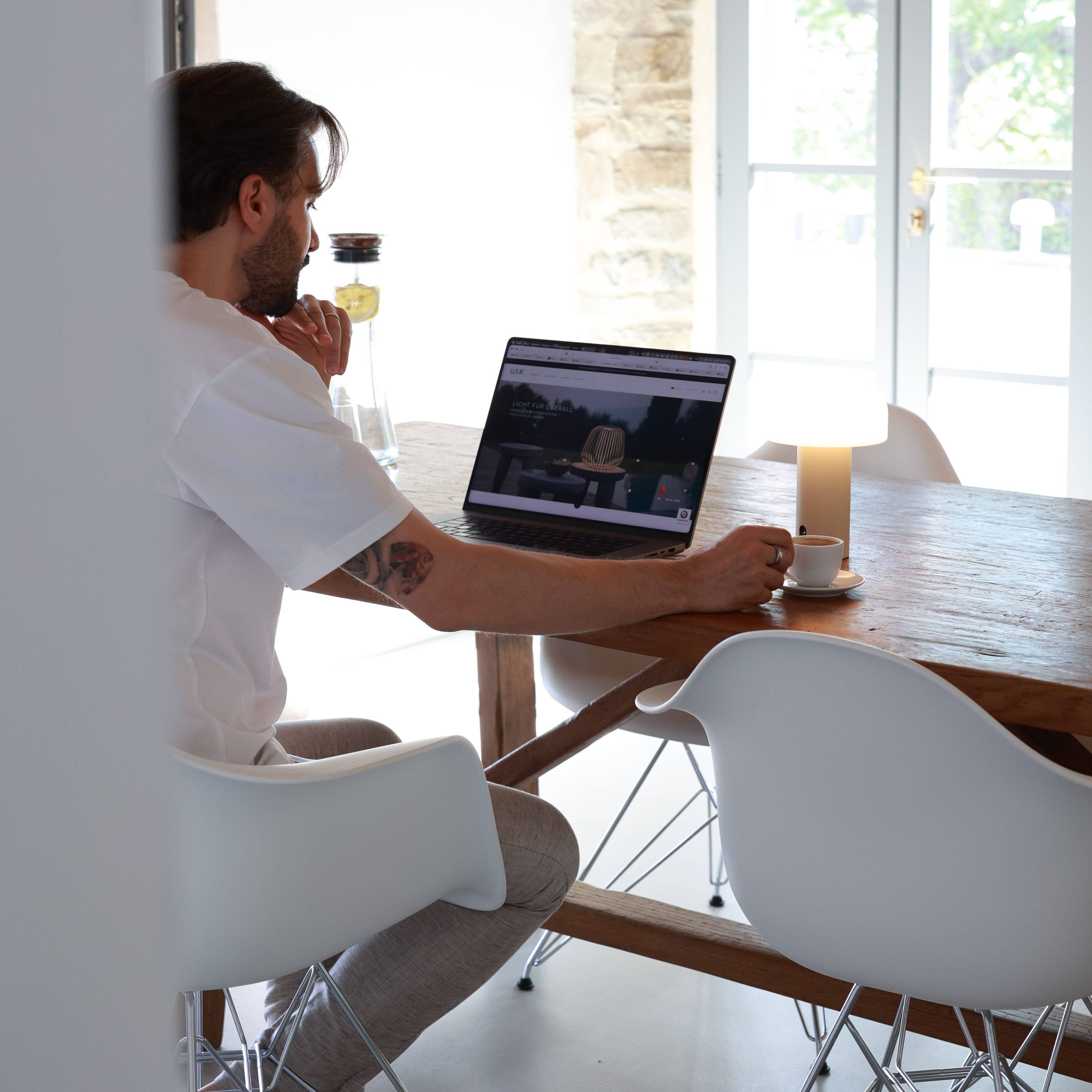 Die akkubetriebne NOA Lampe steht auf einem Esstisch mit einem Mann der am Tisch sitzt, Espresso trinkt und mit dem Laptop arbeitet. 