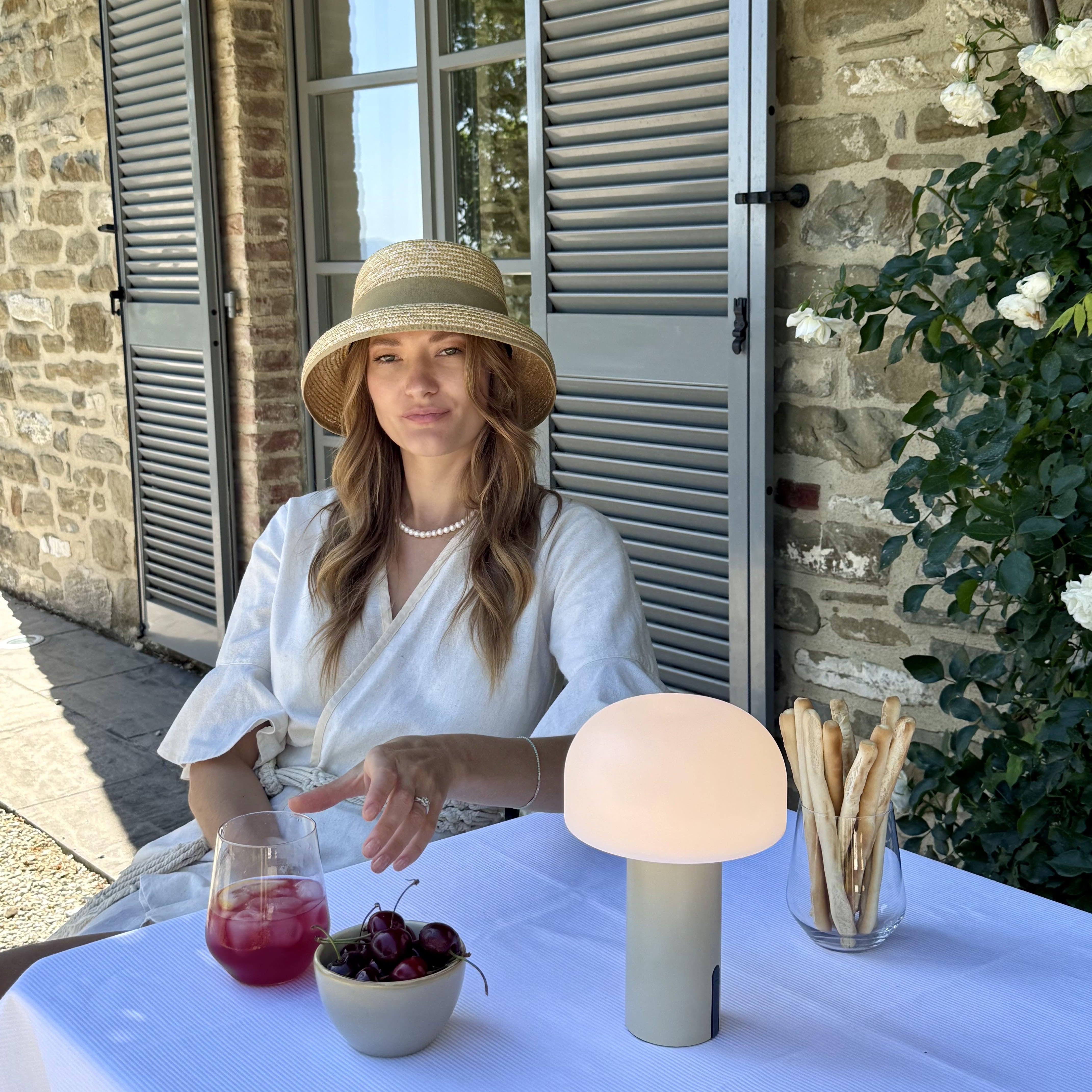 Eine Frau geniest einen kaltes Sommergetraenk und Snacks auf der Terrasse an einem Tisch mit weissem Tischtuch und eienr Noa Lampe auf dem Tisch in einem mediteranen Ambiente