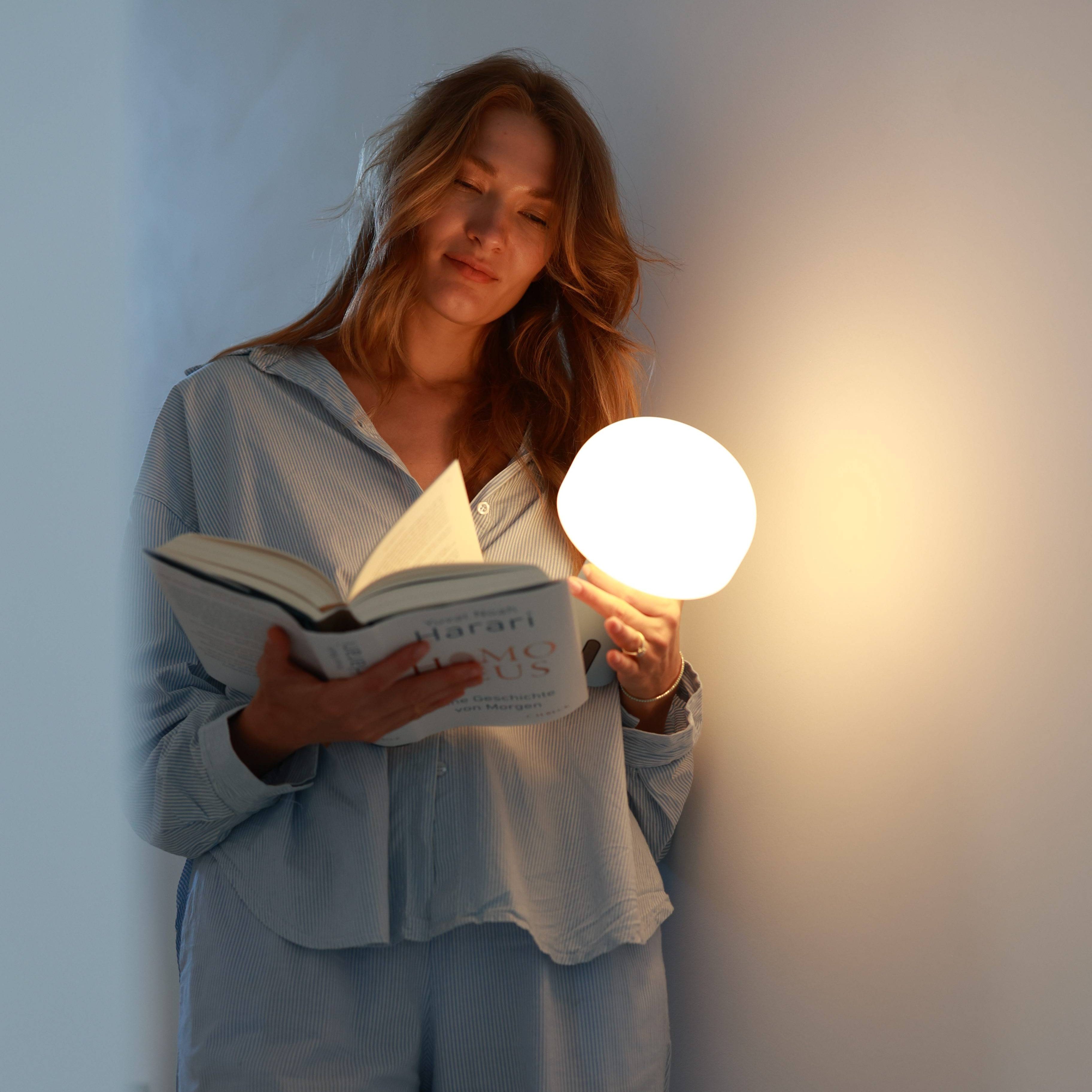 Eine stehende Frau im Schlafanzug hält die kabellose und schlichte NOA Lampe in der Hand und liest ein Buch.