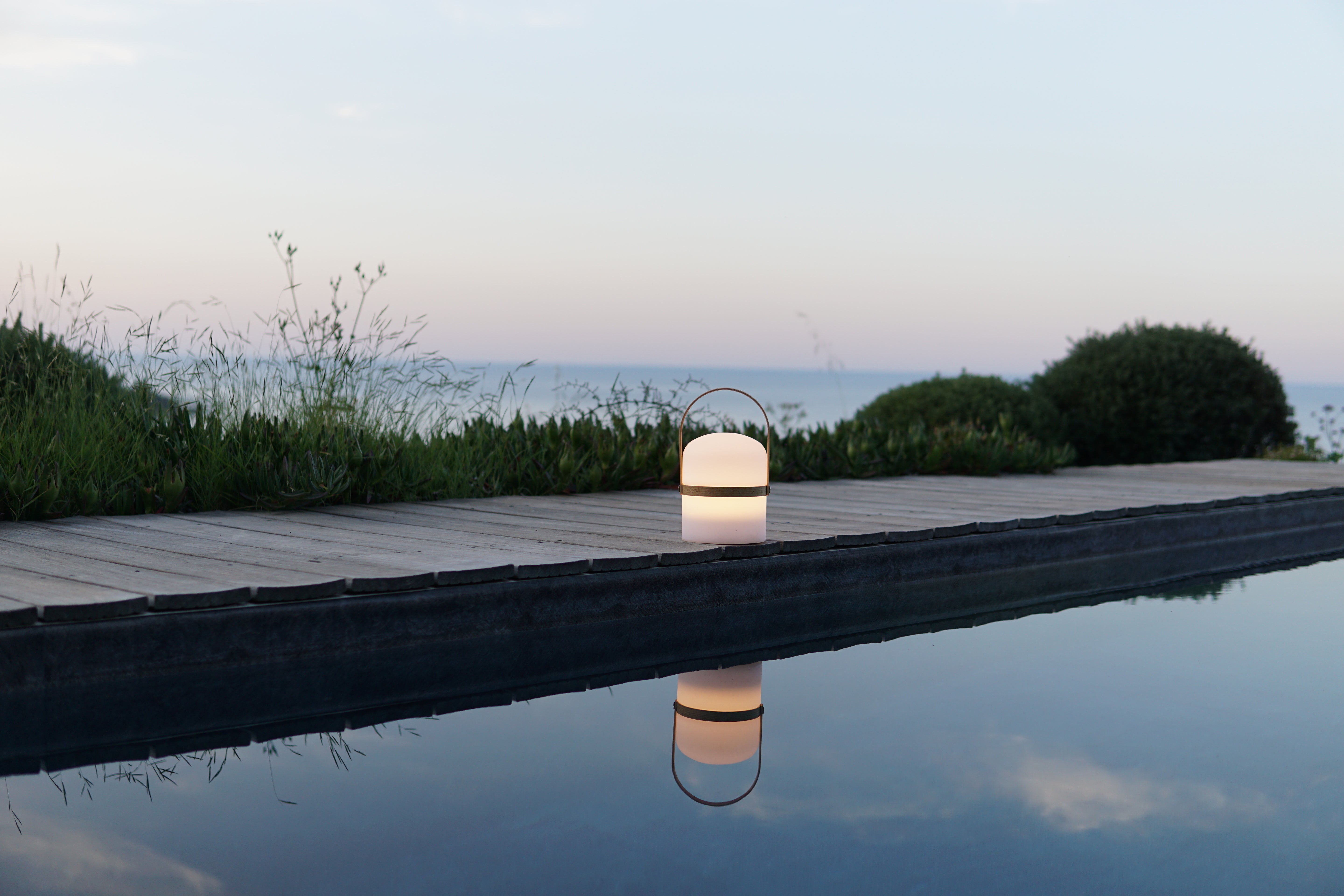 Die Luma Laterne steht in der Abenddämmerung leuchtend auf einer Infinity Poolterrasse von einer Villa, spiegelt sich im Wasser und hat mit Ausblick übers Meer. 
