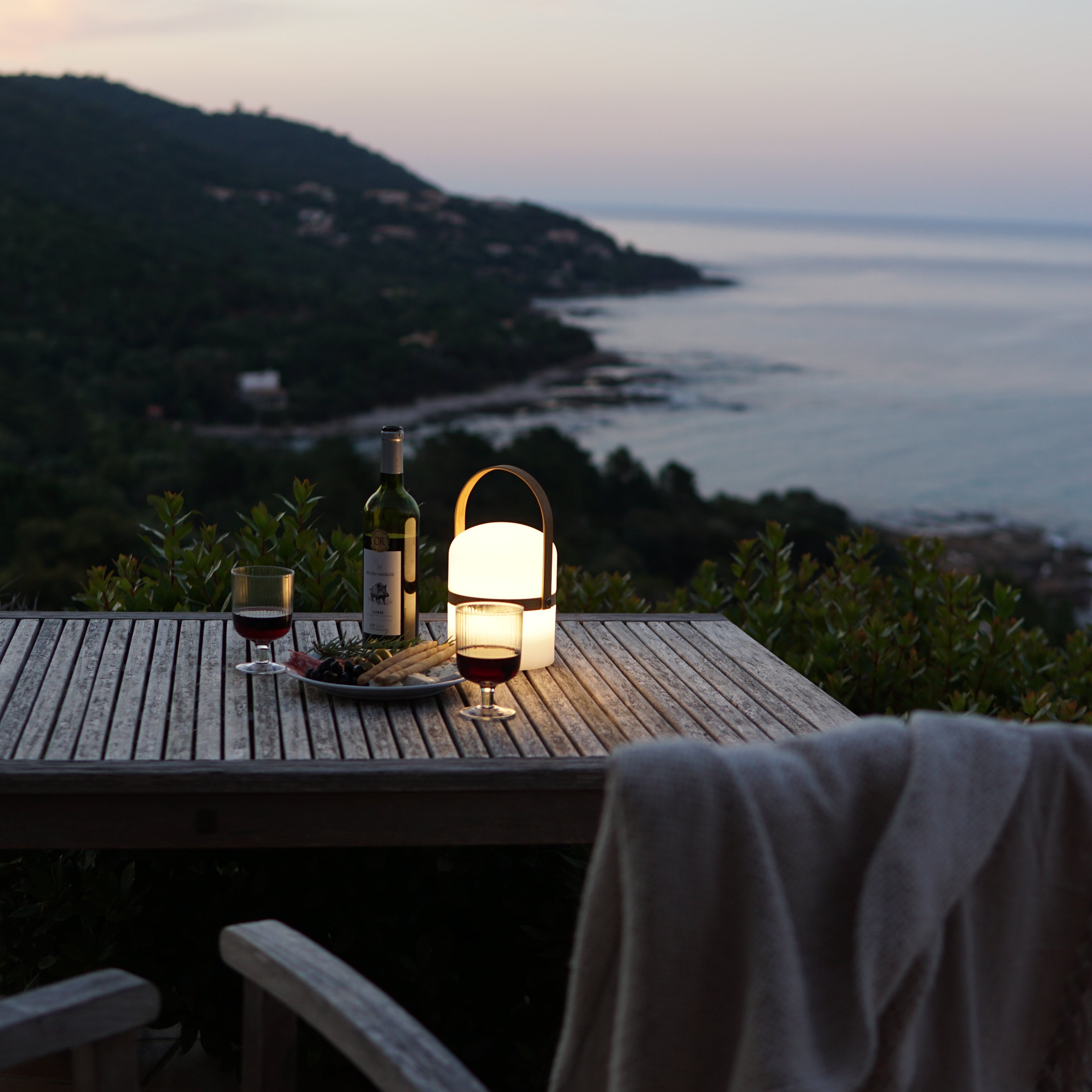 Angeschaltete Luma LED Laterne in der Dämmerung auf Holztisch auf Terrasse in mediterranem Garten, Rotwein und Antipasti mit Ausblich übers Meer. 