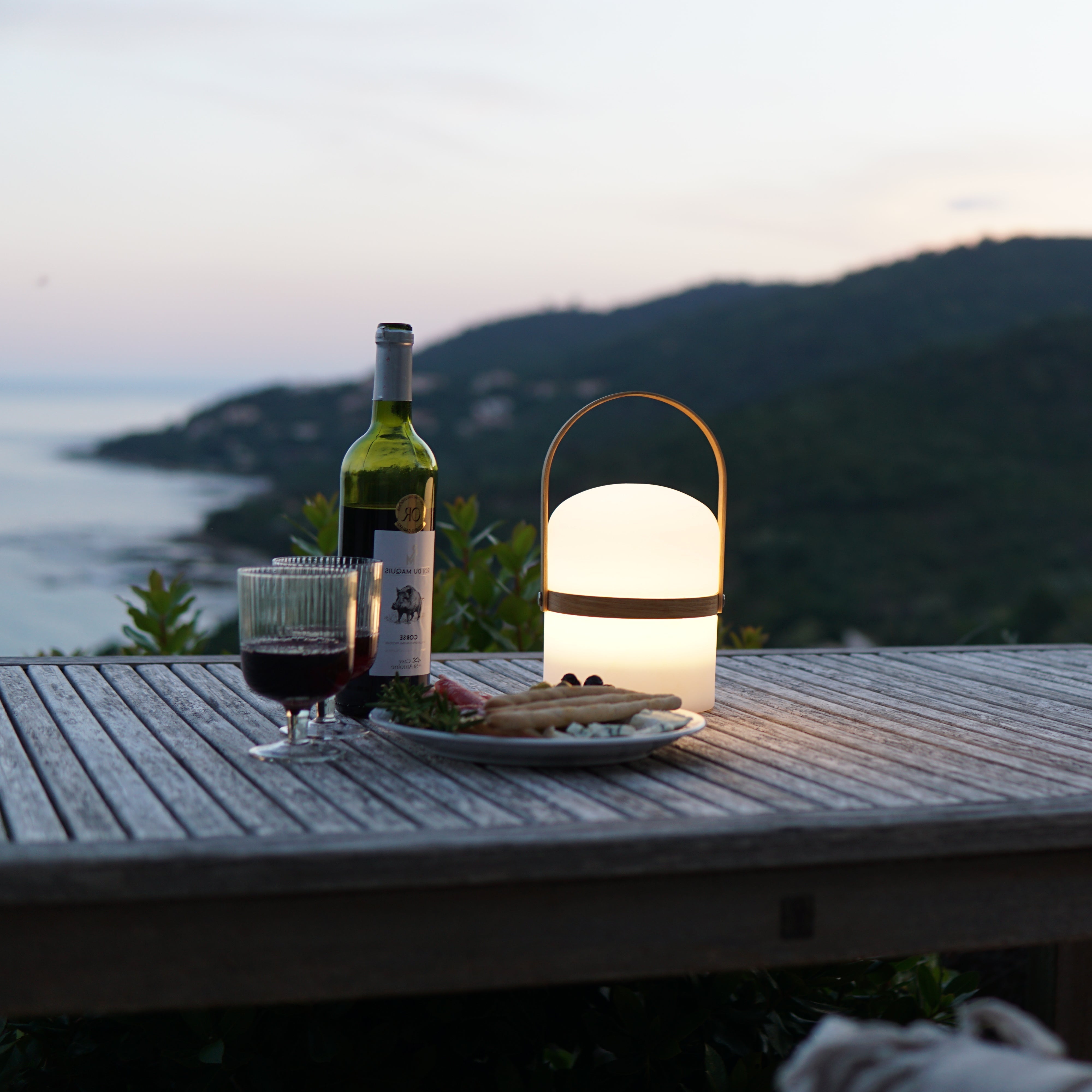 Die gedimmte Luma LED Tischlampe mit  mit Rotwein, Weingläsern und Antipasti auf Terrassentisch und Weitblick übers Meer.