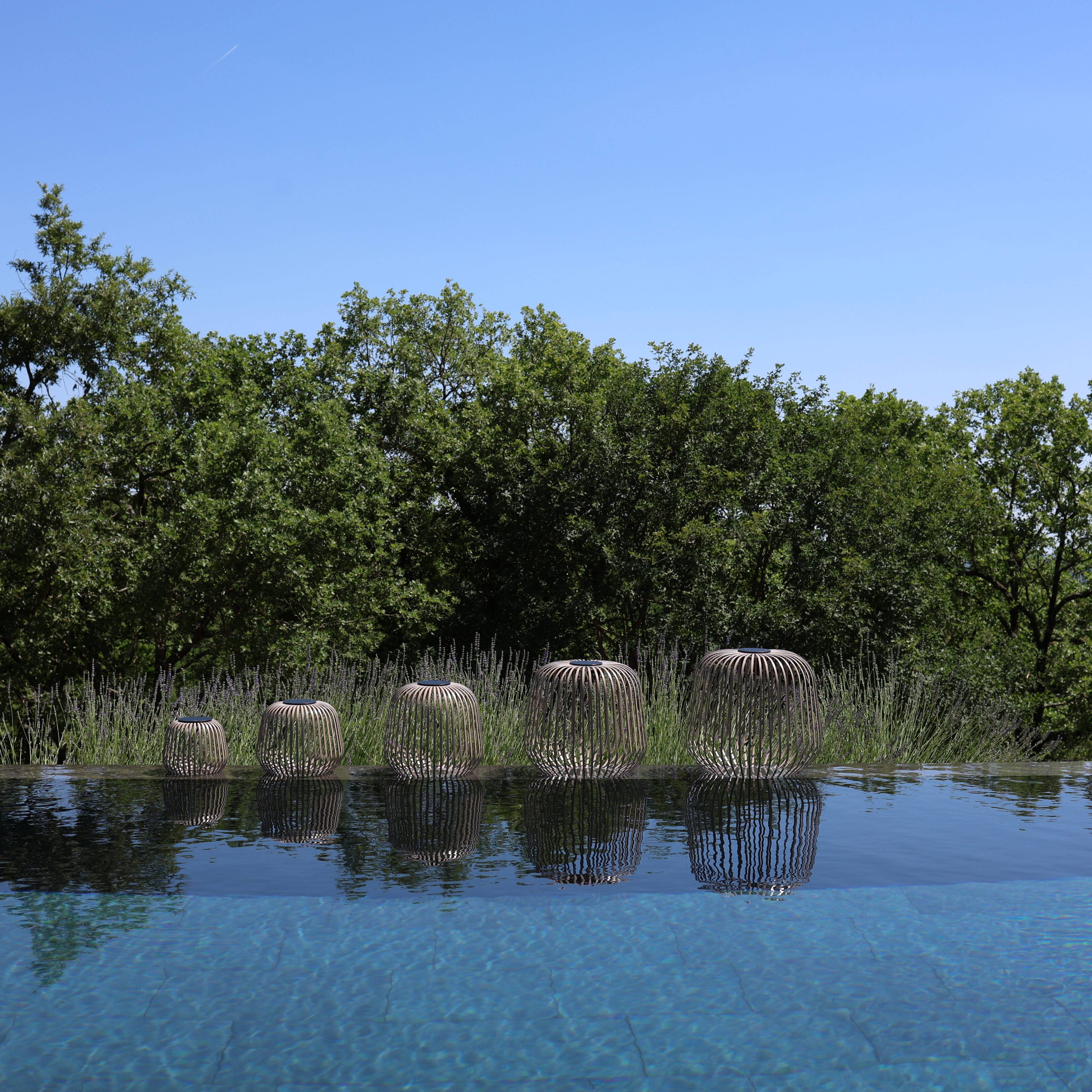 Ein Übersichtbild von allen fünf Größen der Solis Laterne in der Farbe Nomad auf der Kante eines Outdoorpools im Garten vor Lavendel Pflanzen und Bäumen.