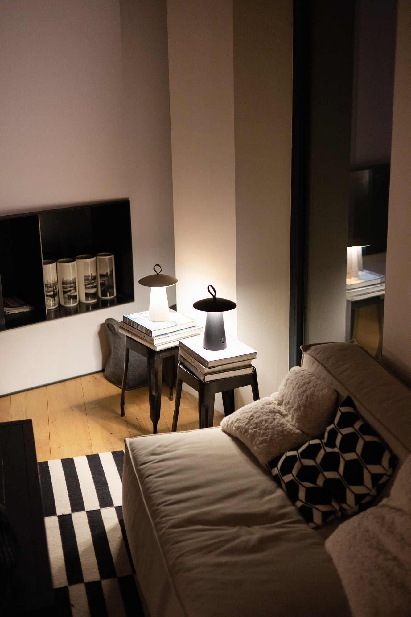 Zwei leuchtende Lyvo Tischleuchten von LUUK Studio in stilvollem Ambiente im dunklen Wohnzimmer auf kleinen Tischen mit warmweißem Lichtkegel. 