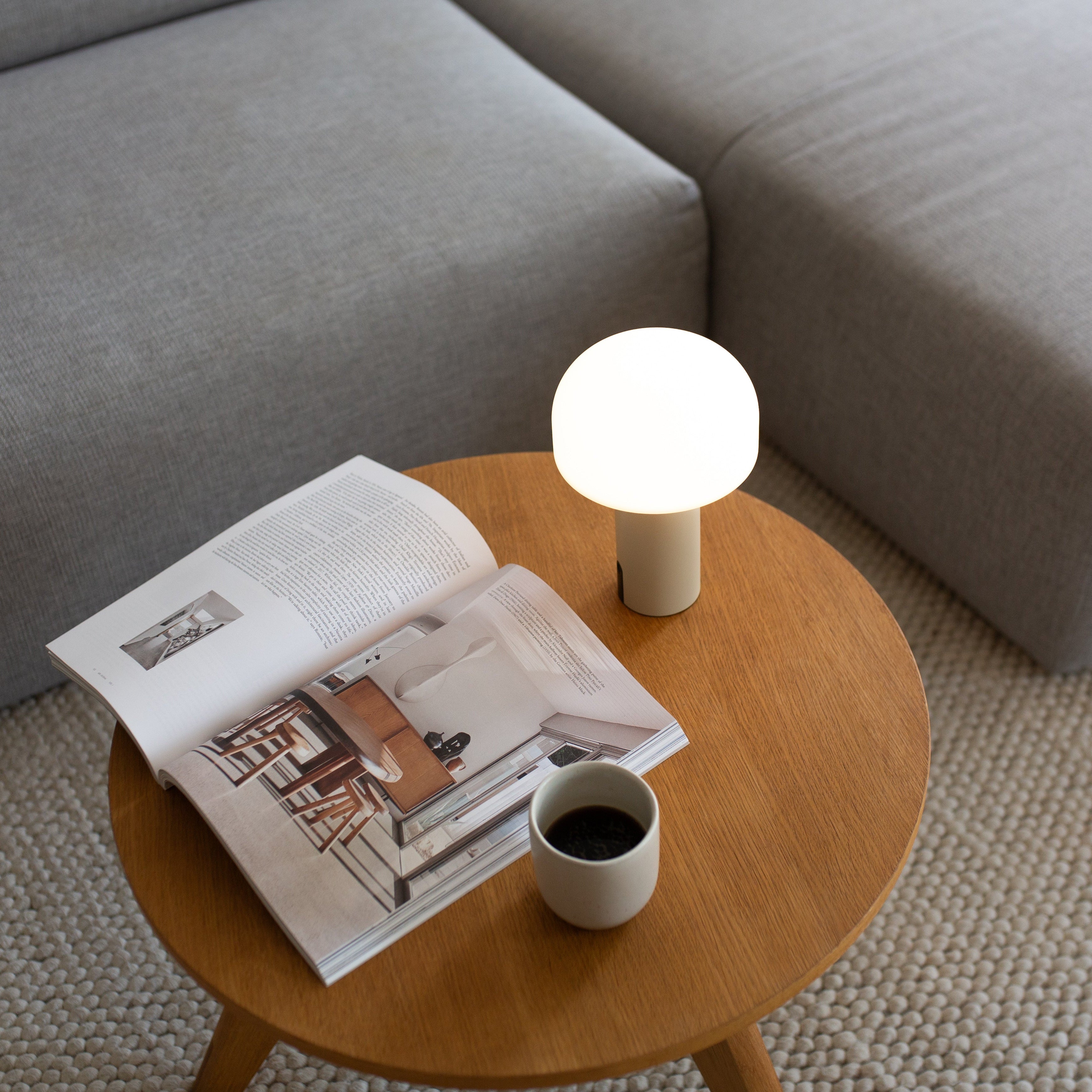 LED-Akku Leuchte Noa in hellem Beige auf einem runden Holztisch im Wohnzimmer, sorgt für sanftes Licht beim Lesen oder Entspannen