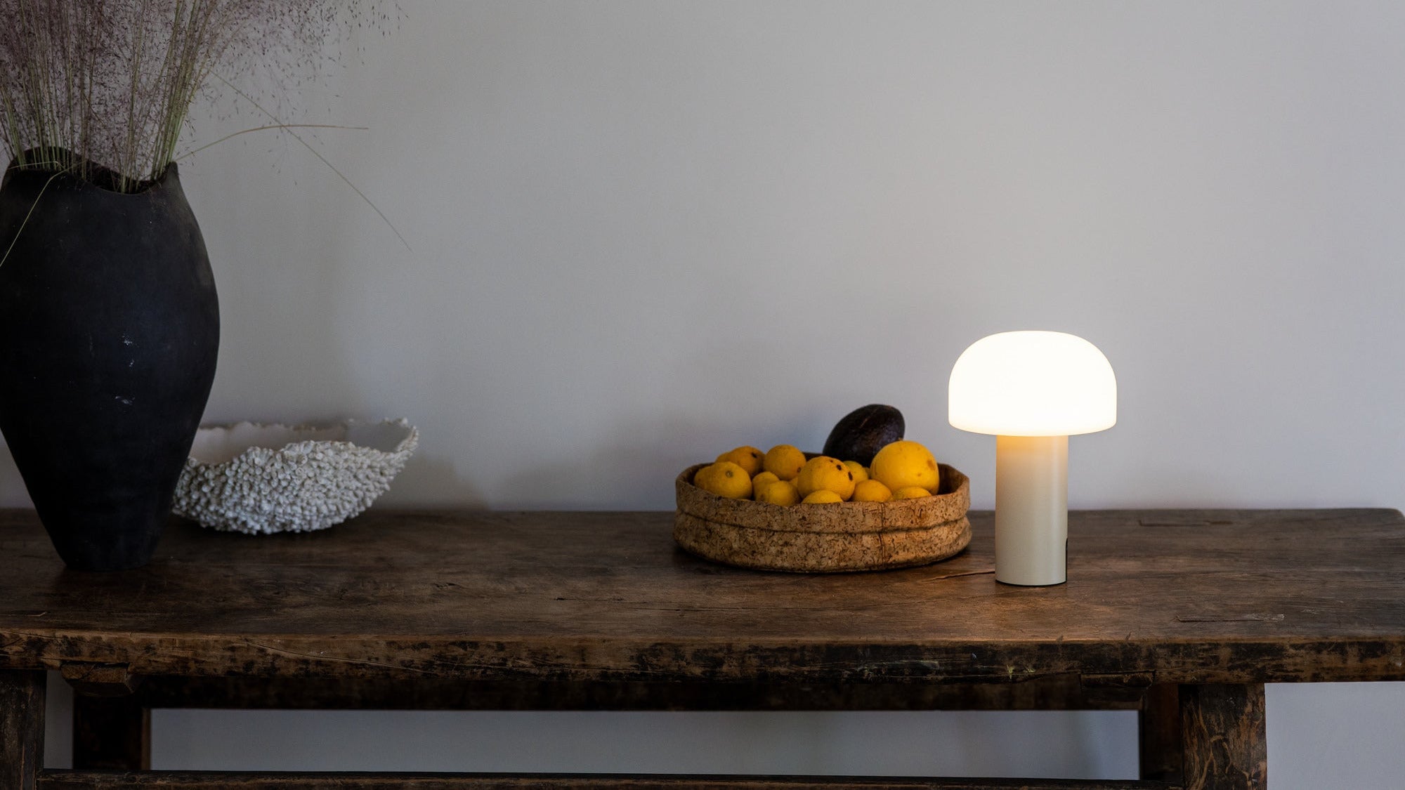 Die LED-Tischleuchte Noa mit pilzförmigem Schirm spendet warmes Licht auf einem rustikalen Holztisch – stimmungsvolle Beleuchtung im minimalistischen Wohnambiente mit natürlichen Materialien.